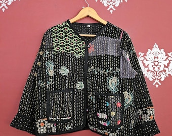 May include: Black patchwork jacket with floral and geometric embroidery. The jacket has a round neckline, two front pockets, and a button-up closure. The jacket is hanging on a wooden hanger against a red wall.