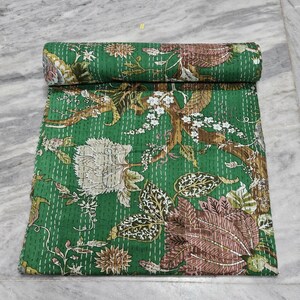 Puede incluir: Un edredón verde enrollado con un estampado floral. El edredón presenta grandes flores estilizadas en tonos marrón, blanco y rosa, sobre un fondo verde. El edredón está cosido a mano con hilo blanco.