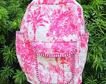 May include: A quilted backpack with a pink and white toile pattern featuring palm trees and zebras. The backpack has a front pocket, side pockets, and a top handle. The edges are trimmed with a pink, black, and white striped fabric.