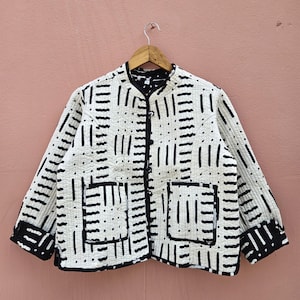 May include: A white quilted jacket with a black geometric pattern. The jacket has a round collar, two front pockets, and a button-up closure. The cuffs and collar are trimmed in black.