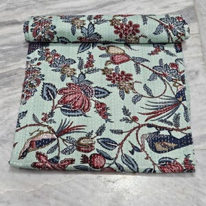 Puede incluir: Un edredón verde claro enrollado con un estampado floral en rojo, azul marino y marrón. El edredón está parcialmente desenrollado, revelando el mismo patrón de flores y hojas. Las costuras son visibles.