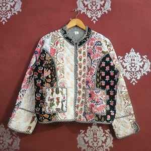Könnte beinhalten: Gesteppte Jacke mit Patchwork-Design mit Blumen- und geometrischen Mustern in Schwarz, Weiß und Rot. Die Jacke hat einen gestreiften Kragen und Besatz sowie zwei Vordertaschen. Sie hängt an einem Holzbügel.