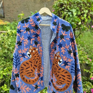 May include: A blue jacket featuring embroidered orange and brown tigers, dark green floral accents, and light pink leafy patterns. The interior lining is visible, displaying a white and navy blue vertical pinstripe fabric pattern.