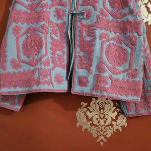 May include: A light blue jacket with intricate pink floral embroidery. The jacket features a detailed pattern of flowers and geometric shapes, with long sleeves and a tie closure. The embroidery is raised, creating a textured effect.