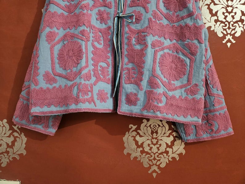 May include: A light blue jacket with intricate pink floral embroidery. The jacket features a detailed pattern of flowers and geometric shapes, with long sleeves and a tie closure. The embroidery is raised, creating a textured effect.