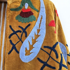 以下が含まれることがあります： マスタード色のベルベットジャケット。青、赤、緑の刺繍が施されており、花や抽象的な形が含まれています。ジャケットの内側の裏地はストライプ柄です。