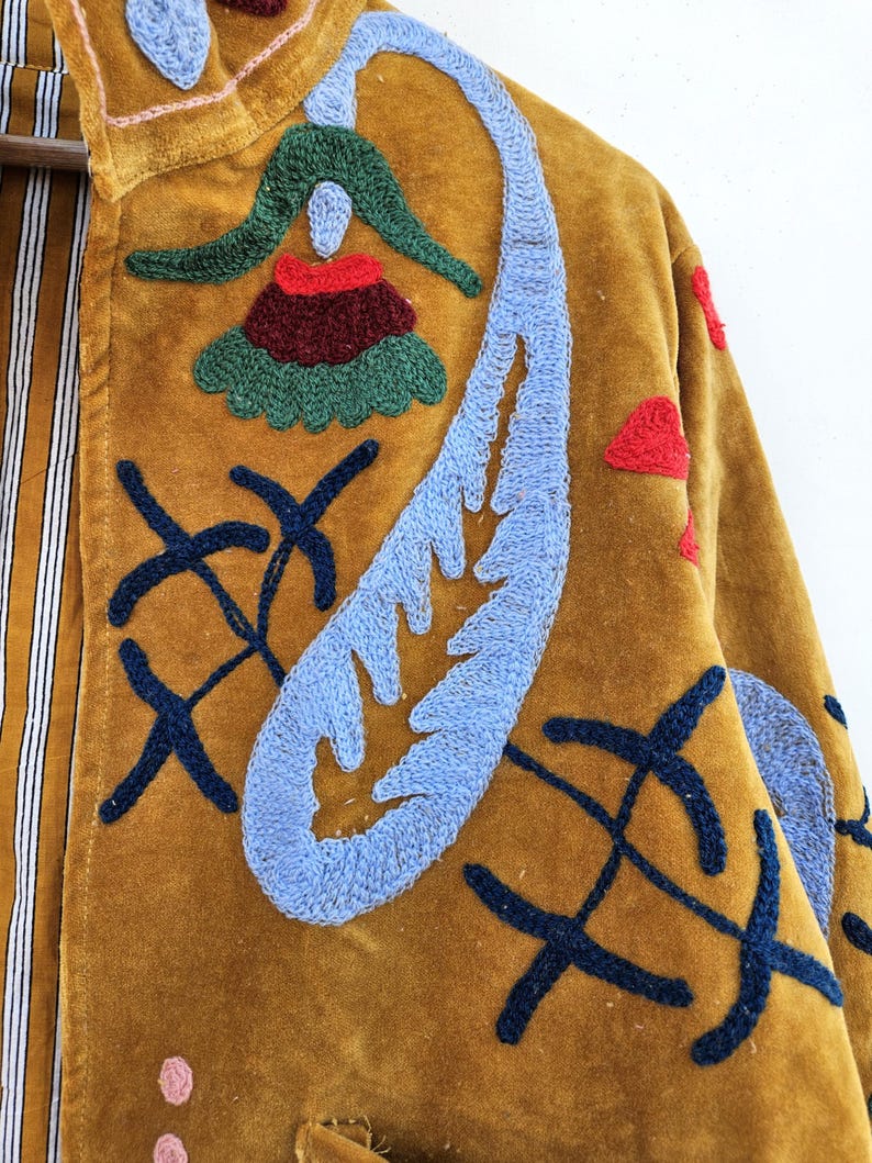 以下が含まれることがあります： マスタード色のベルベットジャケット。青、赤、緑の刺繍が施されており、花や抽象的な形が含まれています。ジャケットの内側の裏地はストライプ柄です。