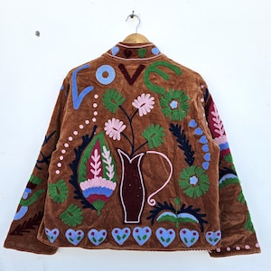 May include: A brown velvet jacket with colourful floral embroidery. The back features a vase with flowers, the word "LOVE", and heart shapes. The embroidery includes blue, green, pink, and red details. The jacket is hanging on a wooden hanger.