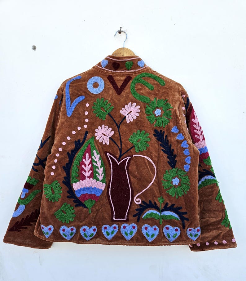 May include: A brown velvet jacket with colourful floral embroidery. The back features a vase with flowers, the word "LOVE", and heart shapes. The embroidery includes blue, green, pink, and red details. The jacket is hanging on a wooden hanger.