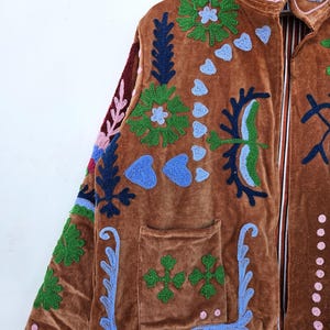 May include: A brown velvet jacket adorned with vibrant embroidered patterns. The jacket showcases green, blue, and pink floral and abstract designs, a front pocket, and a collared neckline. It hangs on a wooden hanger.