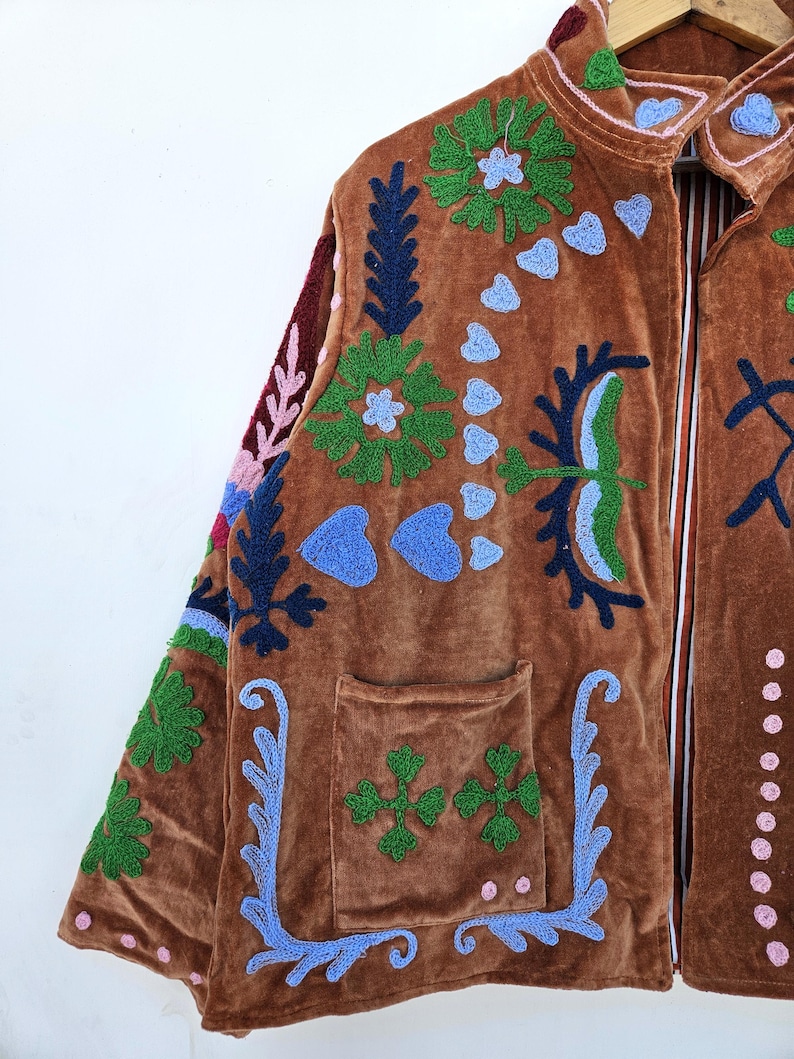 May include: A brown velvet jacket adorned with vibrant embroidered patterns. The jacket showcases green, blue, and pink floral and abstract designs, a front pocket, and a collared neckline. It hangs on a wooden hanger.