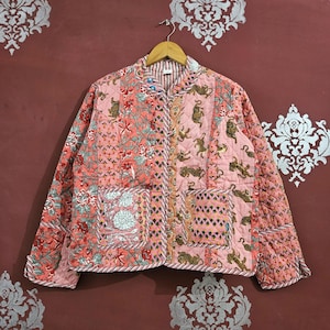 May include: A pink and coral quilted jacket with a patchwork design, showcasing floral and tiger patterns. The jacket has a round collar, long sleeves, and two pockets. It is suspended from a wooden hanger.