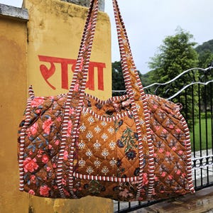 Può includere: Borsa da viaggio trapuntata marrone con motivi floreali e geometrici. La borsa presenta un motivo ripetuto di rose rosa e foglie blu. I manici e i bordi sono rifiniti con un design a righe. La borsa è sospesa davanti a un muro giallo.