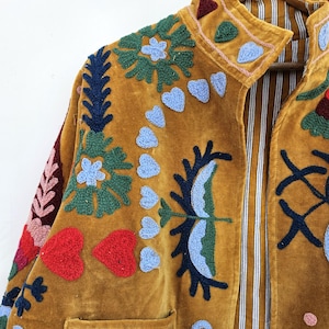 以下が含まれることがあります： マスタード色のベルベットジャケット、複雑な刺繍が施されています。 ジャケットは、青、緑、赤の色合いの花柄とハート型のデザインが特徴です。 襟とポケットが見え、裏地はストライプ柄です。