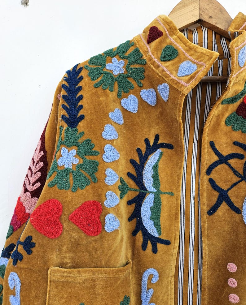 以下が含まれることがあります： マスタード色のベルベットジャケット、複雑な刺繍が施されています。 ジャケットは、青、緑、赤の色合いの花柄とハート型のデザインが特徴です。 襟とポケットが見え、裏地はストライプ柄です。