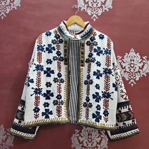 Peut inclure: Veste blanche ornée de broderies florales bleues et de tiges marron. La veste a un col montant et un panneau rayé sur le devant. Les manches et le col sont ornés de broderies dorées et bleues. La veste est suspendue à un cintre en bois.