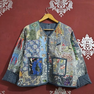 Può includere: Giacca patchwork con scollo a V e due tasche frontali. La giacca presenta una varietà di motivi floreali e geometrici nei toni del blu, grigio e rosso. La giacca è appesa a un'appendiabiti di legno.