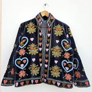 Könnte beinhalten: Dunkelblaue Samtjacke mit farbenfrohen Stickereien. Die Jacke zeigt Blumen- und Herzmotive in Orange, Rosa, Blau und Grün. Sie hängt an einem Holzkleiderbügel.