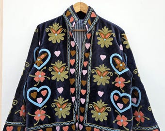 May include: A dark blue velvet jacket with colorful embroidery. The jacket features floral and heart designs in shades of orange, pink, blue, and green. The jacket is hanging on a wooden hanger.