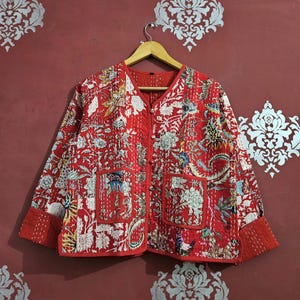 May include: Red quilted jacket with a floral pattern, featuring a V-neck, two front pockets, and button closures. The jacket has red trim and cuffs. The floral design includes white, blue, and yellow accents. The jacket is hanging on a wooden hanger.