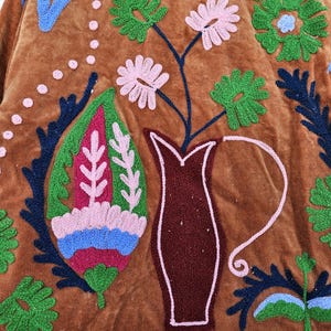 May include: Close-up of a brown velvet textile with colourful embroidered floral and geometric designs. The embroidery features pink, green, blue, and white thread, creating a textured and decorative pattern. A vase with flowers is also embroidered.