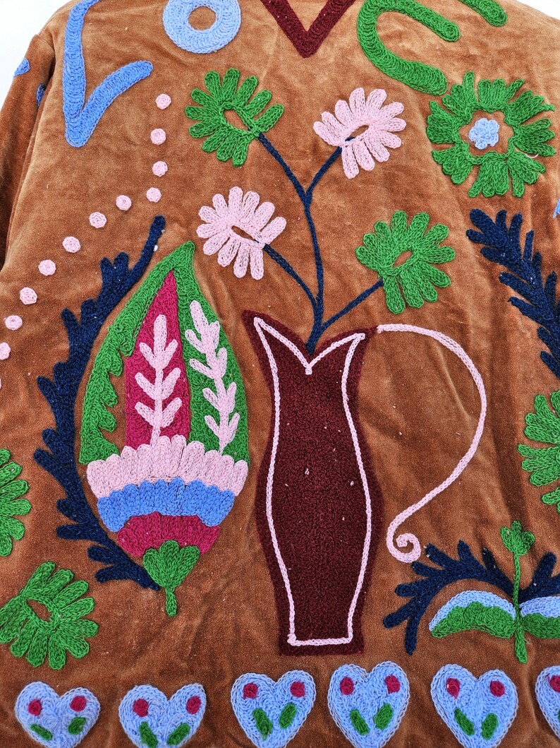 May include: Close-up of a brown velvet textile with colourful embroidered floral and geometric designs. The embroidery features pink, green, blue, and white thread, creating a textured and decorative pattern. A vase with flowers is also embroidered.
