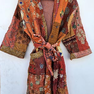 Op de afbeelding: Een patchwork badjas in tinten oranje, rood en bruin, met ingewikkelde bloemen- en geometrische patronen. De badjas heeft een gestreepte voering en een bijpassende strikriem. De badjas hangt aan een houten hanger.