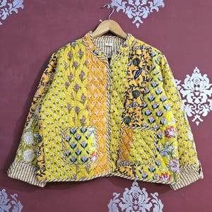 以下が含まれることがあります： 黄色を基調としたキルティングジャケット。花柄と幾何学模様が特徴です。スタンドカラー、2つのポケット、装飾ボタンが付いています。裏地とトリムはストライプ柄です。木製のハンガーに掛けられています。