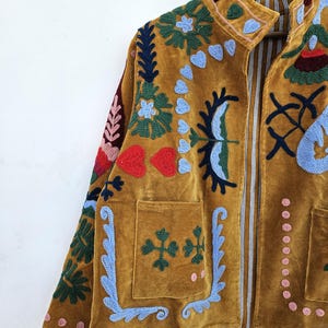 以下が含まれることがあります： マスタード色のベルベットジャケット、カラフルな花の刺繍入り。 ジャケットはハイカラー、2つのフロントポケット、ストライプの裏地が特徴です。 刺繍には青、緑、赤、ピンクのアクセントが含まれており、ボヘミアンな美学を作り出しています。