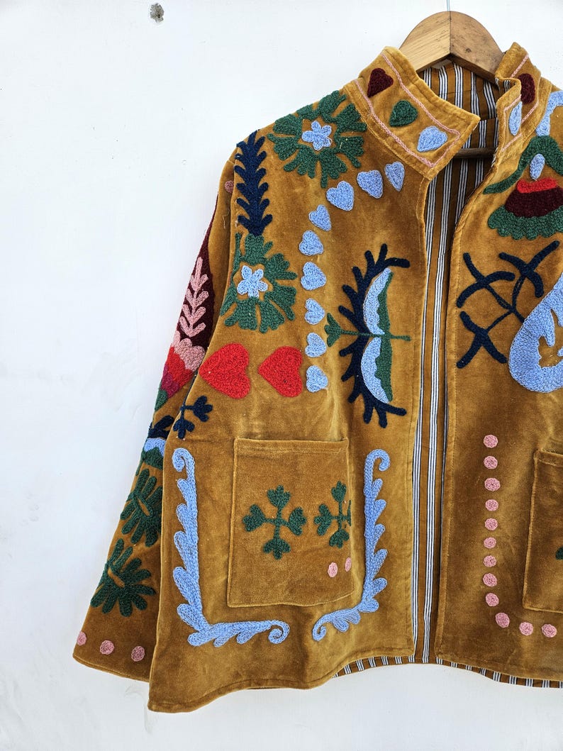 以下が含まれることがあります： マスタード色のベルベットジャケット、カラフルな花の刺繍入り。 ジャケットはハイカラー、2つのフロントポケット、ストライプの裏地が特徴です。 刺繍には青、緑、赤、ピンクのアクセントが含まれており、ボヘミアンな美学を作り出しています。