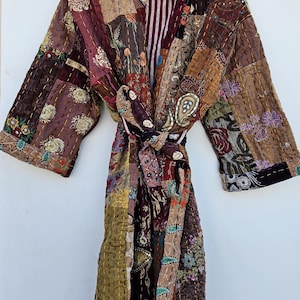 Puede incluir: Bata de patchwork con cinturón anudado, con una variedad de telas en tonos marrón, burdeos y dorado. La bata está colgada de una percha de madera sobre un fondo blanco. La bata tiene un forro a rayas.