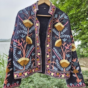 Puede incluir: Chaqueta azul marino con bordados florales coloridos. La chaqueta abierta, con cuello alto, está adornada con flores y hojas bordadas en marrón, rosa, amarillo y azul. Cuelga de una percha de madera sobre un fondo natural.