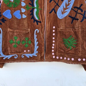 May include: Brown velvet jacket with embroidered floral and abstract designs in blue, green, and pink. Features two front pockets and a white zipper. The jacket's design includes a variety of shapes and patterns.
