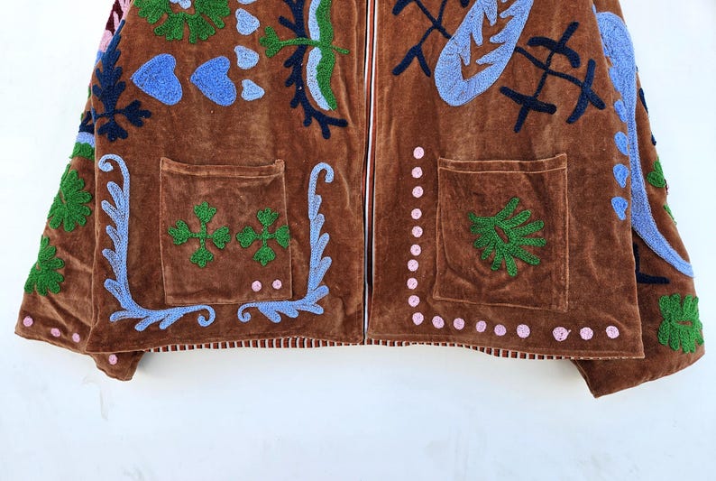 May include: Brown velvet jacket with embroidered floral and abstract designs in blue, green, and pink. Features two front pockets and a white zipper. The jacket's design includes a variety of shapes and patterns.