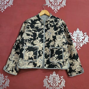 Peut inclure: Veste matelassée à motifs floraux noirs et beiges, col officier et deux poches avant. La veste a une doublure et une bordure rayées, et est suspendue à un cintre en bois. La veste mesure environ 63,5 cm de long.