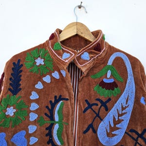May include: A brown velvet jacket with colourful floral embroidery. The jacket features a high collar and a full-length zip. The embroidery includes green, blue, and maroon floral and paisley designs. The jacket is hanging on a wooden hanger.