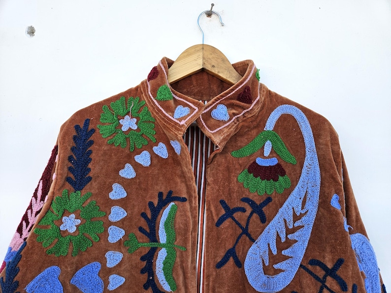 May include: A brown velvet jacket with colourful floral embroidery. The jacket features a high collar and a full-length zip. The embroidery includes green, blue, and maroon floral and paisley designs. The jacket is hanging on a wooden hanger.