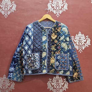 May include: A quilted jacket with a patchwork design in shades of blue, featuring floral and paisley patterns. The jacket has two front pockets and a striped lining. The jacket is displayed on a wooden hanger.