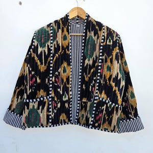 Peut inclure: Veste matelassée noire avec un motif ikat coloré dans les tons de vert, or et rouge. La veste a une doublure et des poignets rayés, et est suspendue à un cintre en bois.