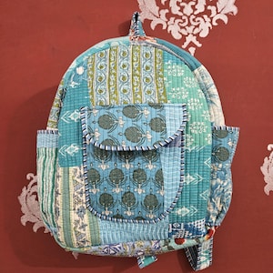 May include: A quilted backpack in shades of blue and green, featuring various patterned fabric patches. The backpack has a front pocket and side pockets, with a handle at the top. The stitching is visible, and the overall design is patchwork.