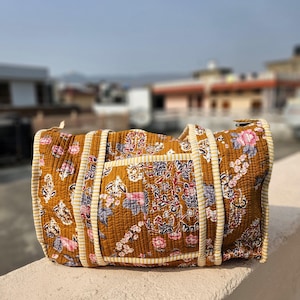 Puede incluir: Una bolsa de viaje acolchada con un estampado floral en tonos marrón, rosa y azul. La bolsa tiene dos asas y un ribete a rayas. La bolsa mide aproximadamente 46 cm de largo.