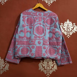 May include: A light blue jacket with intricate pink floral embroidery. The jacket features long sleeves and a round neckline. The embroidery creates a textured, raised pattern across the garment, showcasing a detailed floral design.