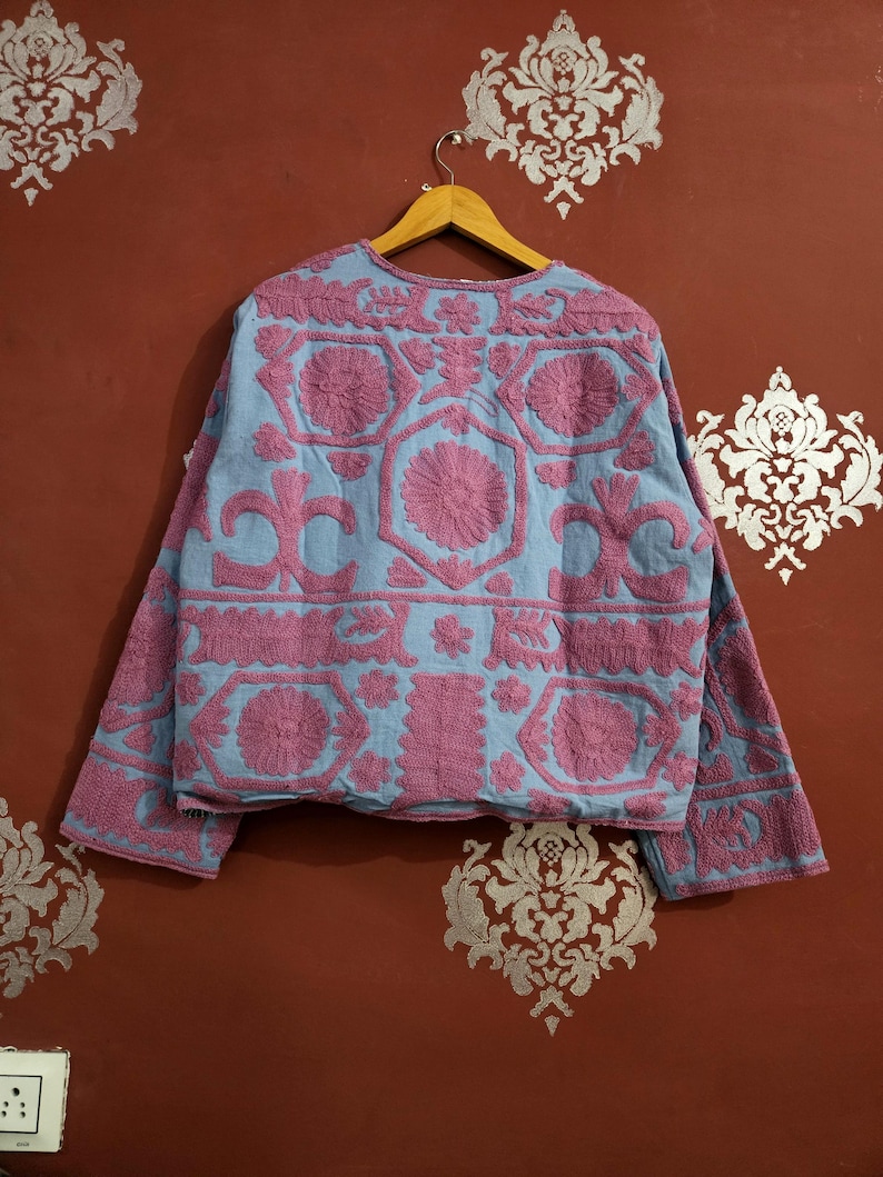 May include: A light blue jacket with intricate pink floral embroidery. The jacket features long sleeves and a round neckline. The embroidery creates a textured, raised pattern across the garment, showcasing a detailed floral design.