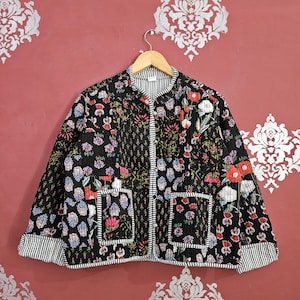 May include: Black quilted jacket adorned with a colourful floral design in red, pink, blue, and white. The jacket has a striped collar, cuffs, and pocket trim. It features two front pockets and a button-up closure.