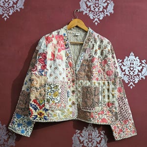Peut inclure: Veste patchwork avec divers tissus à motifs floraux dans les tons de rose, rouge, bleu et crème. La veste a un col en V, deux poches et est suspendue à un cintre en bois.