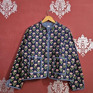 May include: A navy blue quilted jacket with a floral pattern of pink flowers and green leaves. The jacket has a striped lining and trim, two front pockets, and a button-up closure. The jacket is hanging on a wooden hanger.