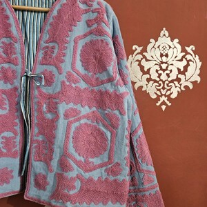 May include: A light blue jacket with intricate pink embroidery. The jacket features a V-neck and long sleeves. The interior lining has a blue and white striped pattern. The jacket is hanging on a wooden hanger.