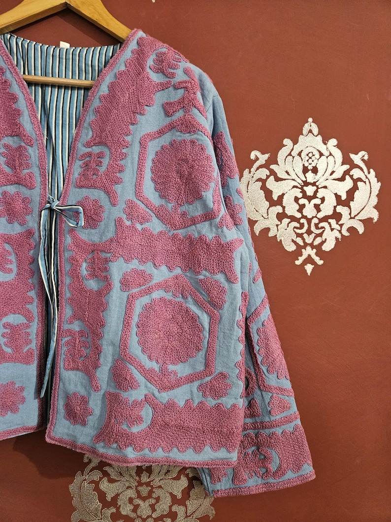 May include: A light blue jacket with intricate pink embroidery. The jacket features a V-neck and long sleeves. The interior lining has a blue and white striped pattern. The jacket is hanging on a wooden hanger.