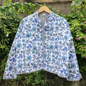 May include: A quilted jacket with a floral design in blue and green on a white base. The jacket features a mandarin collar, two front pockets, and black and white striped trim. It is displayed on a wooden hanger.