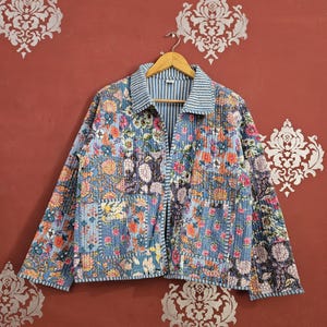 Puede incluir: Chaqueta acolchada con diseño floral de patchwork. La chaqueta presenta una base azul claro con varios estampados florales en tonos naranja, rosa y morado. El cuello y el ribete tienen un estampado de rayas azules y blancas. La chaqueta está colgada de una percha de madera.